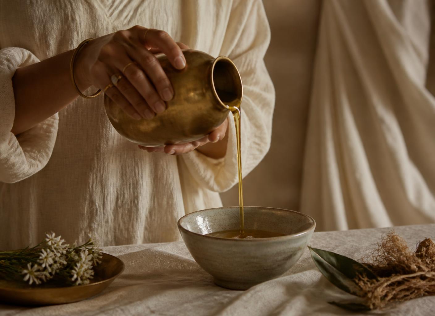 Golden oil being poured from a brass vessel into a ceramic bowl